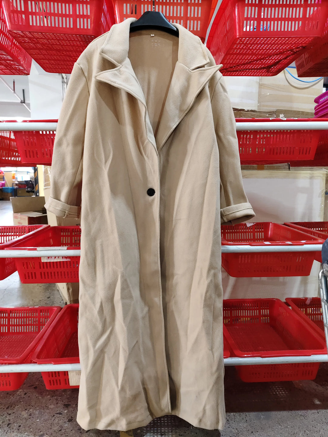 Beige long coat with lapel collar and black button hanging on a rack in a warehouse with red plastic baskets