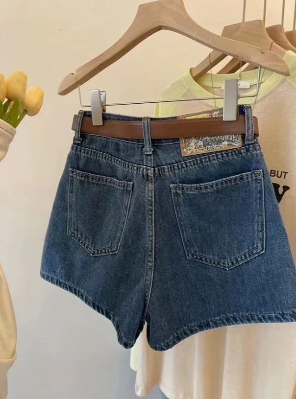 Dark denim high-waisted shorts with brown belt hanging on wooden hanger against light background