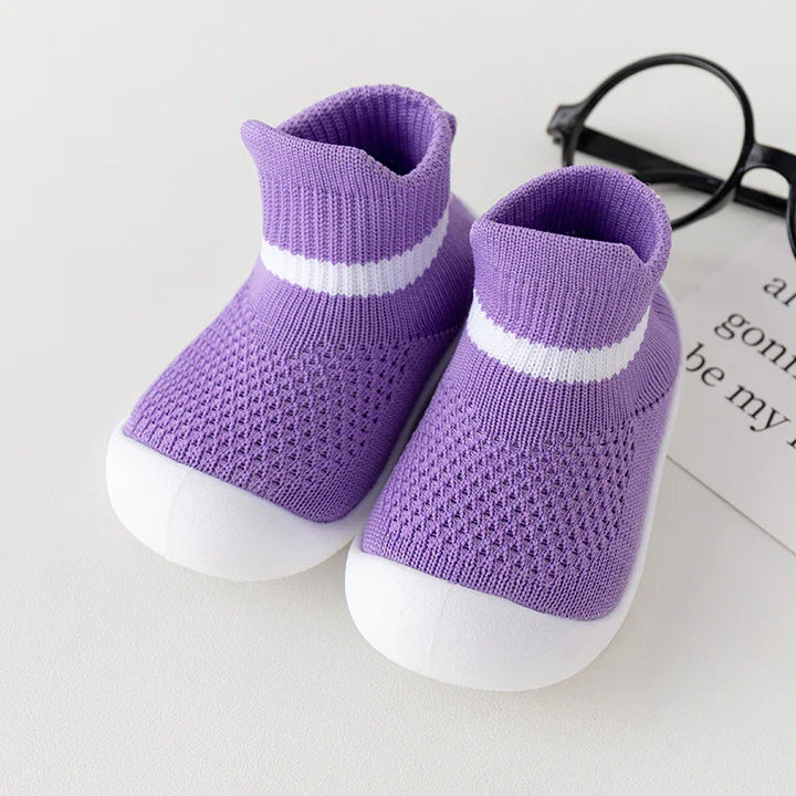 Pair of purple baby sock shoes with white soles and stripe, placed on white surface with black glasses nearby
