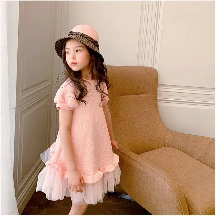 Little girl in pink textured dress and beige-brown hat standing by tan armchair in elegant room
