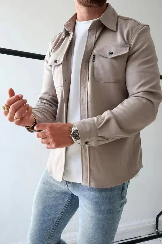 man wearing beige button-up shirt over white t-shirt with light blue jeans, accessorized with watch and rings