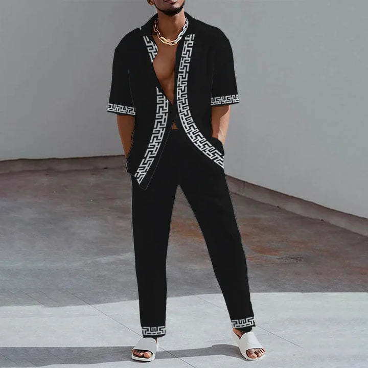 Man wearing black short-sleeve shirt and pants with white Greek key pattern, white sandals, and gold chain necklace