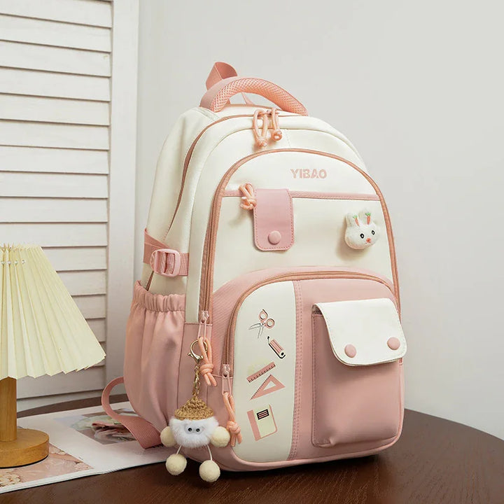 Pink and white YIBAO school backpack with cute bunny and pompom keychains on wooden table