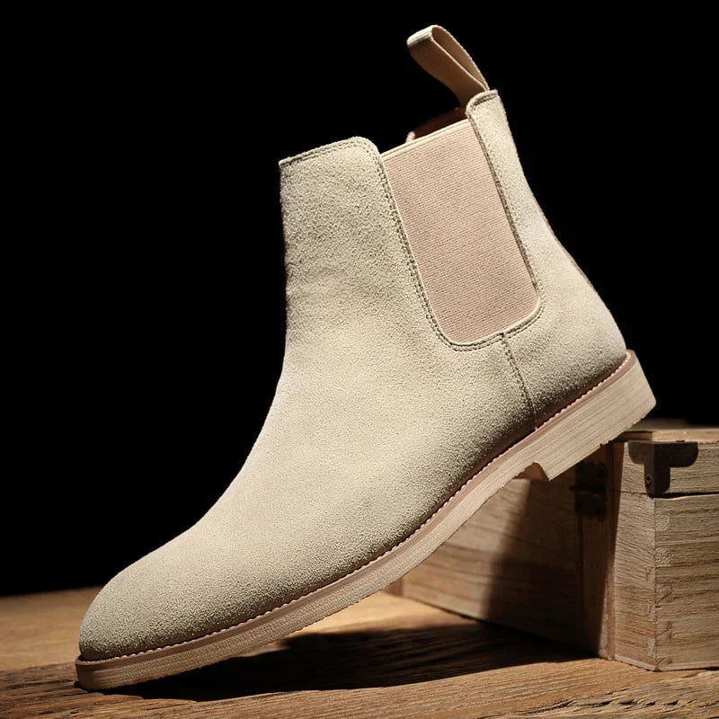 Beige suede Chelsea boot with elastic side panel and pull tab on wooden surface against black background