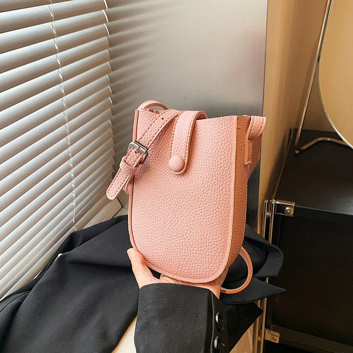 Hand holding small textured pink leather crossbody bag with adjustable strap near window blinds