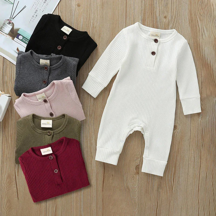 Ribbed cotton baby rompers in white, black, gray, pink, olive, and maroon with buttons on wooden floor