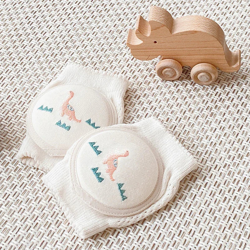 Soft white baby knee pads with cute dinosaur embroidery next to wooden toy rhino on textured mat