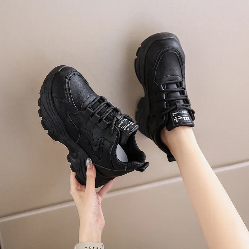 Black chunky platform sneakers with lace-up design worn on feet against beige background
