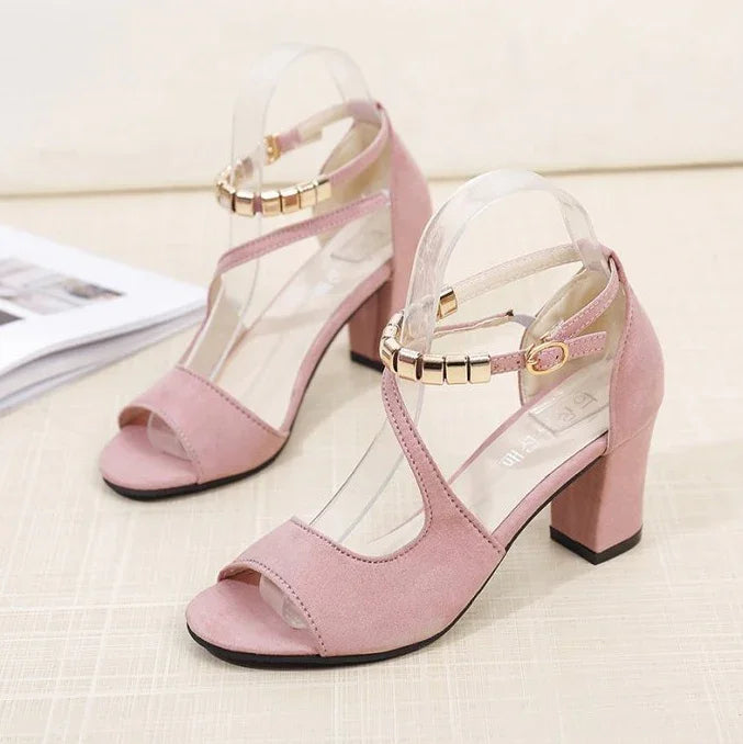 Pair of pink suede block heel sandals with ankle straps and gold buckle accents on beige surface