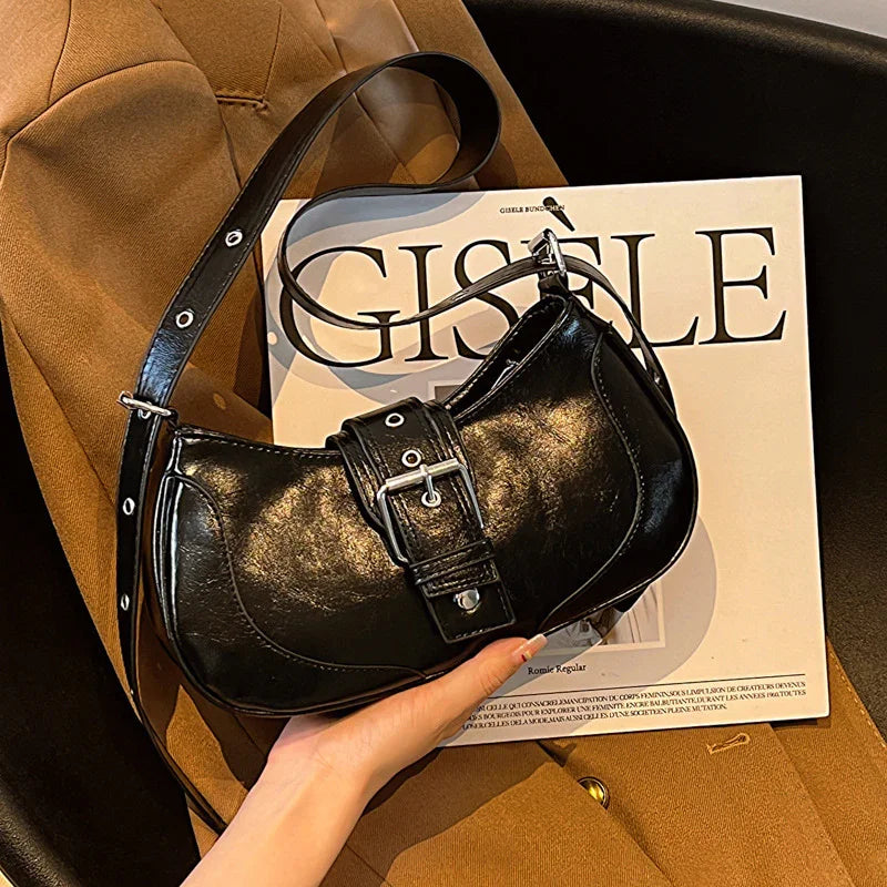 Hand holding black leather shoulder bag with buckle, placed on brown jacket and Gisele magazine