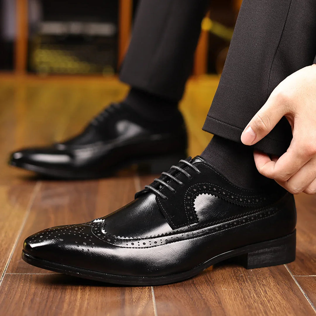 Black leather men's dress wingtip shoes with brogue detailing worn indoors on wooden floor