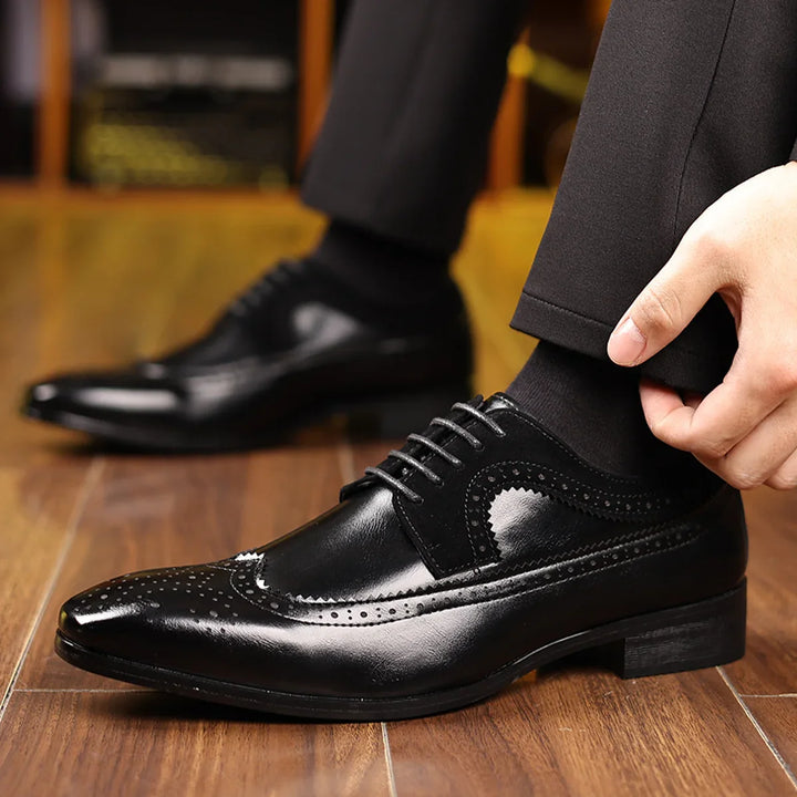 Black leather men's dress wingtip shoes with brogue detailing worn indoors on wooden floor