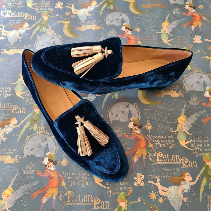 Pair of navy blue velvet loafers with gold tassels on a Peter Pan themed background