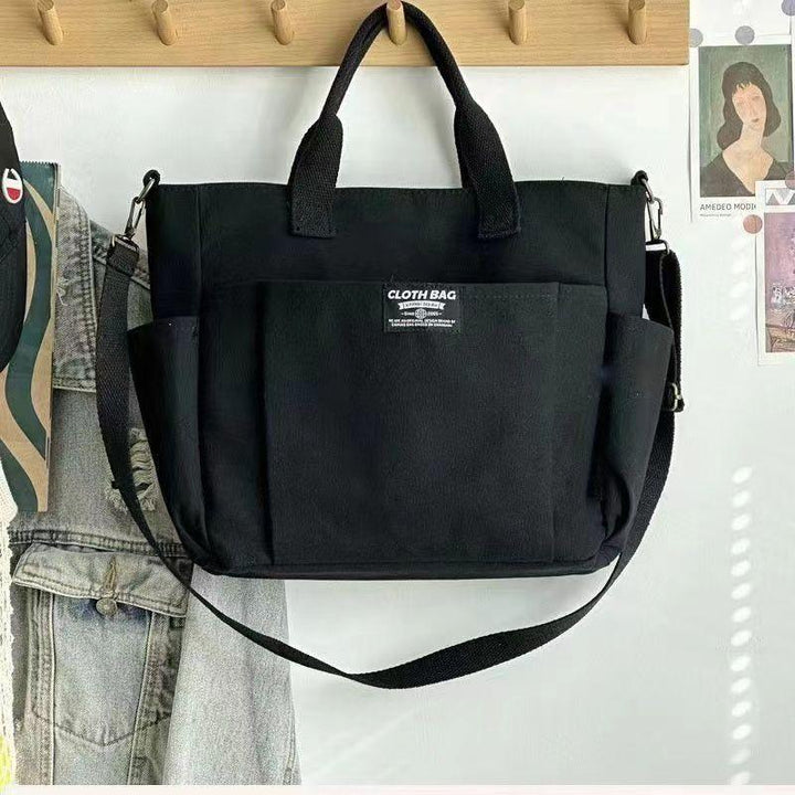 Black cloth tote bag with front pocket and shoulder strap hanging on wooden hooks with denim jacket