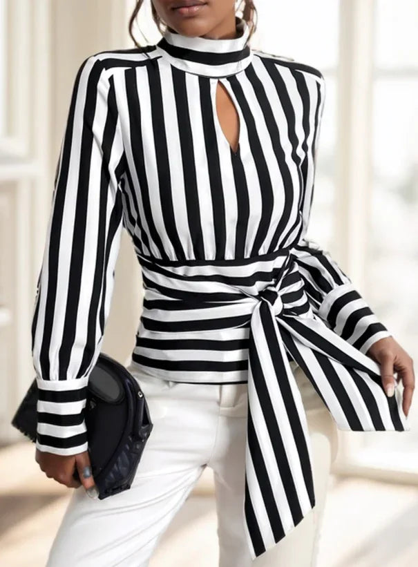 Woman wearing black and white vertical striped long sleeve keyhole blouse with waist tie and white pants, holding black clutch