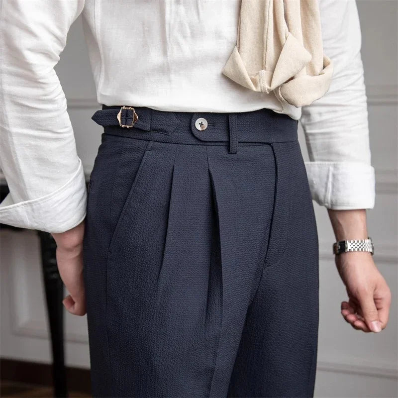 Man wearing navy pleated trousers with side adjuster and white shirt, with beige sweater draped over shoulder