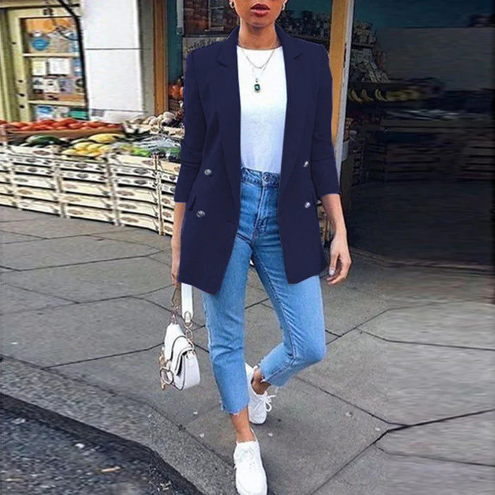Woman wearing navy blazer, white top, light blue jeans, and white sneakers on city sidewalk with market stalls