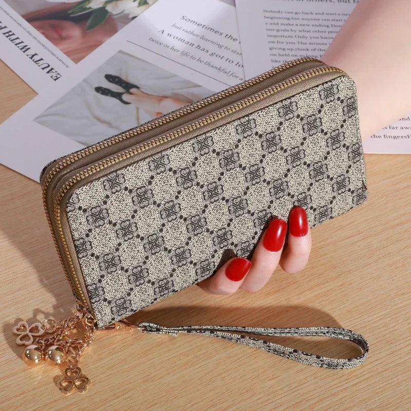 Patterned beige and black zippered wallet with wrist strap and gold heart and bell charms held by hand with red nail polish on wooden surface