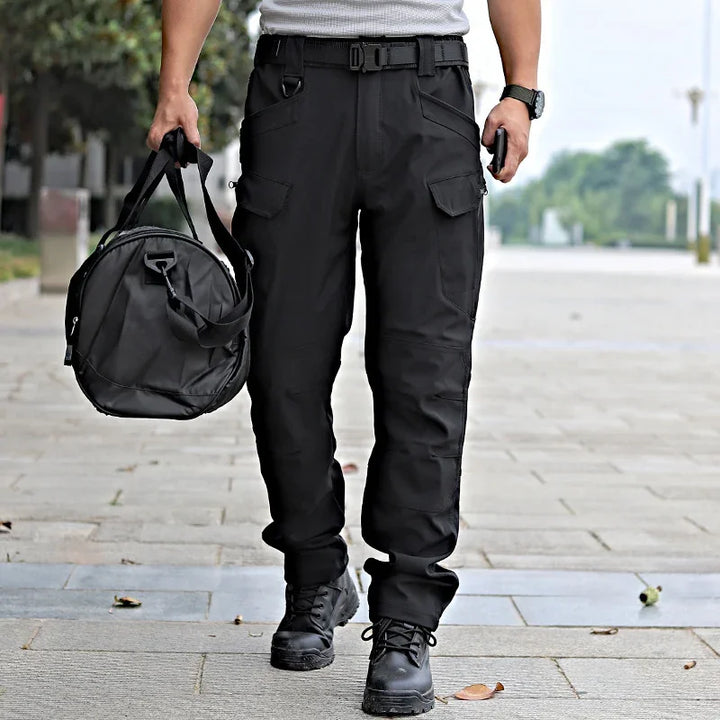 Man wearing black tactical cargo pants with belt and black boots, carrying black duffel bag outdoors