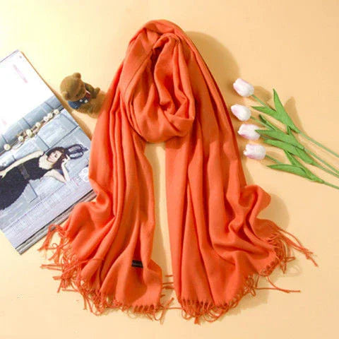 Bright orange fringed scarf displayed with white tulips, a teddy bear, and a fashion magazine on light background