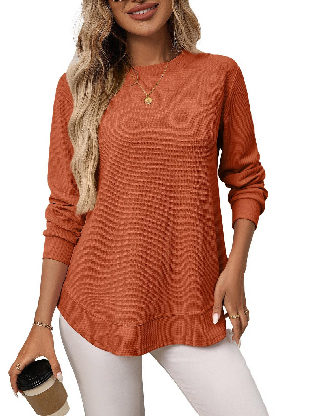 Woman wearing rust orange long sleeve waffle knit top with white pants, holding coffee cup