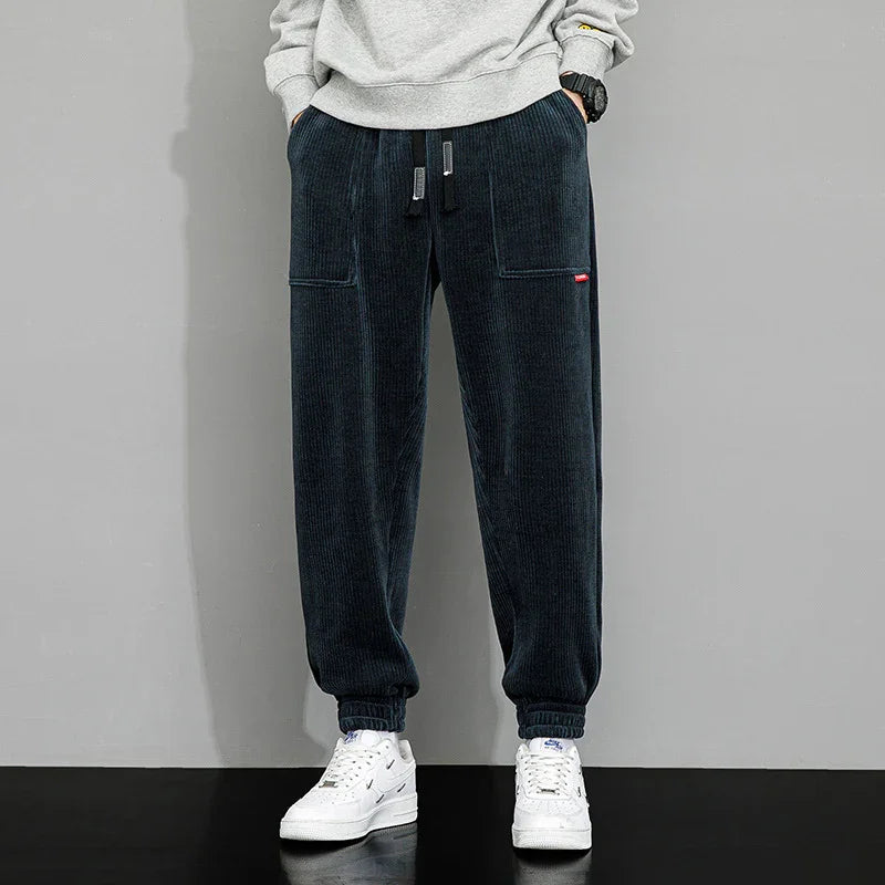 Man wearing black corduroy jogger pants with front pockets and white sneakers against gray wall