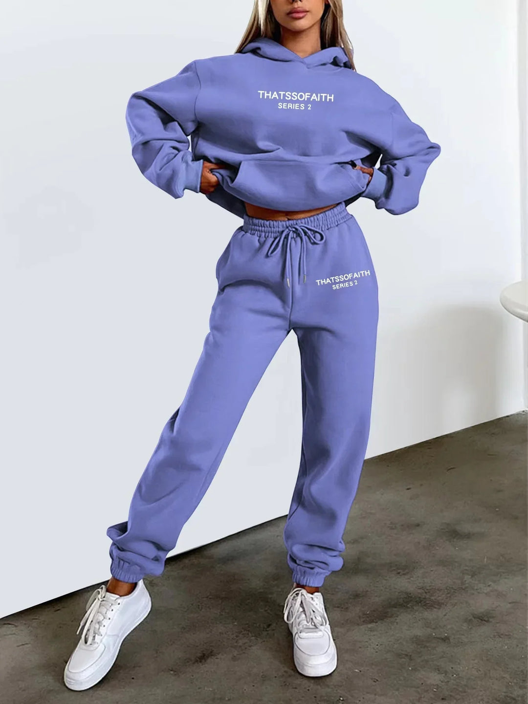 Woman wearing purple Thatssofaith Series 2 hoodie and joggers with white sneakers on concrete floor