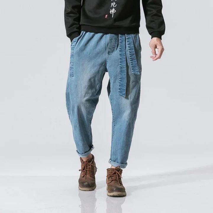 man wearing loose fit light blue denim jogger pants with belt and brown boots on gray background