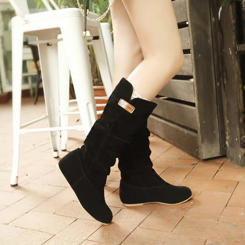 Black suede mid-calf women's boots with wedge heels worn indoors on tiled floor near white furniture