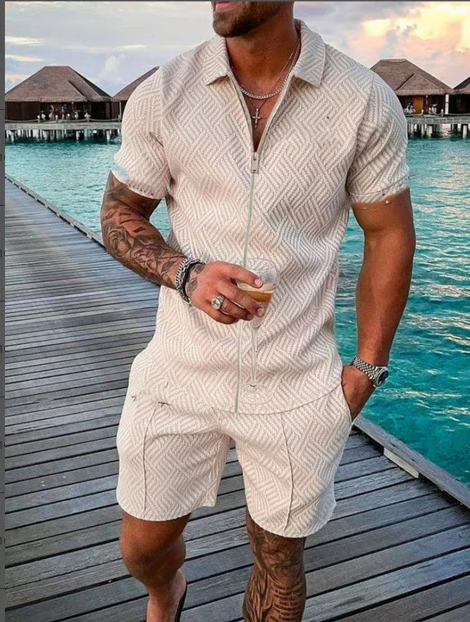 Man with tattoos wearing beige patterned matching short-sleeve shirt and shorts, holding a drink on wooden pier by turquoise water and huts