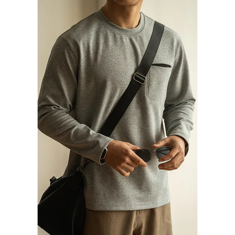 Man wearing gray long sleeve shirt with chest pocket, holding sunglasses and black crossbody bag