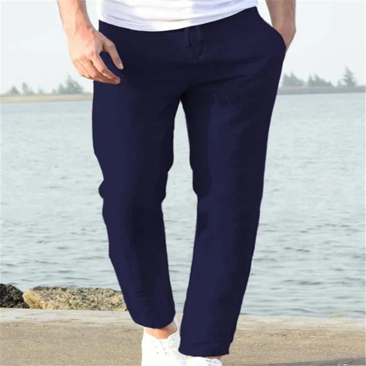 Man wearing navy blue casual pants and white sneakers standing by waterfront with distant trees