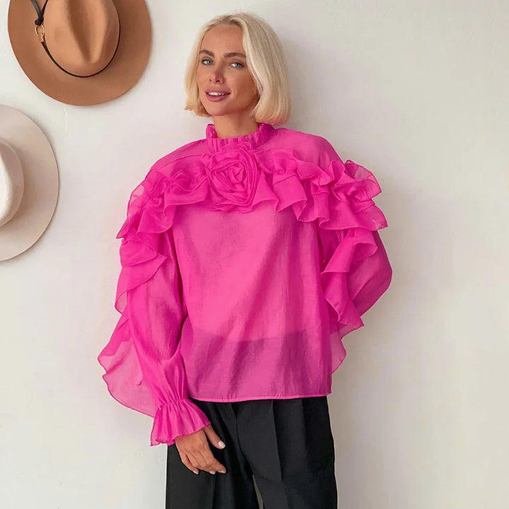 Blonde woman wearing sheer pink ruffled blouse with flower detail, standing against white wall with hanging hats