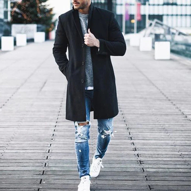 Man wearing black coat, gray sweater, ripped blue jeans, and white sneakers walking on wooden boardwalk