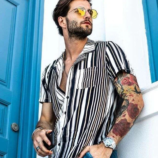 stylish man with tattooed arm wearing black and white striped short-sleeve shirt and yellow sunglasses by blue door