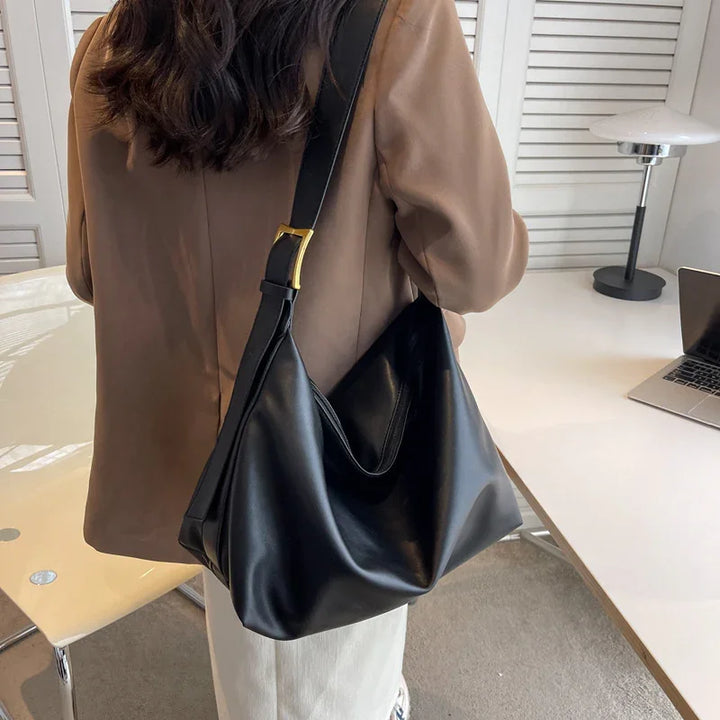 Woman wearing beige blazer carrying large black leather shoulder bag indoors at white desk with laptop