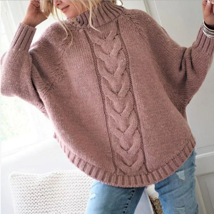 Woman wearing oversized dusty pink cable knit turtleneck sweater with batwing sleeves and ripped blue jeans indoors