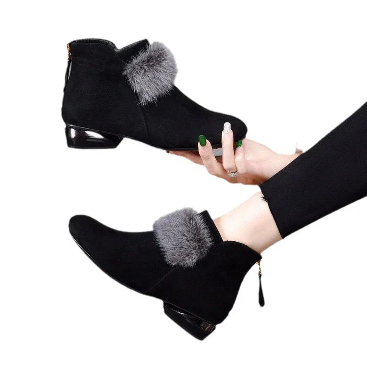 Black suede ankle boots with gray fur trim and low glossy heel, worn on feet and held by hand