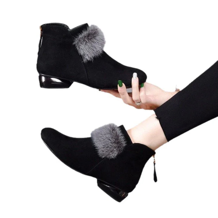 Black suede ankle boots with gray fur trim and low glossy heel, worn on feet and held by hand
