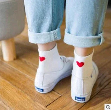 Person wearing white Keds sneakers and white ankle socks with red heart designs indoors on wooden floor