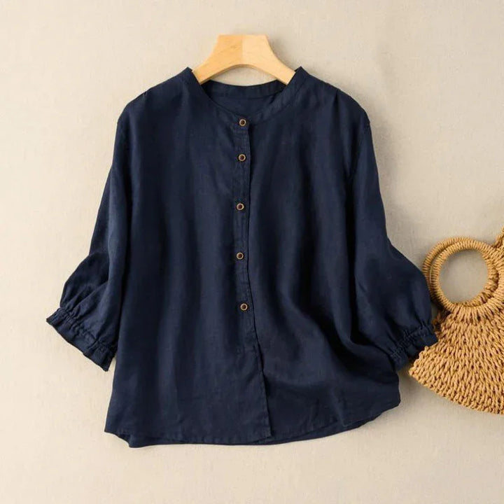 Navy blue button-up blouse with three-quarter sleeves on wooden hanger beside a woven rattan handbag