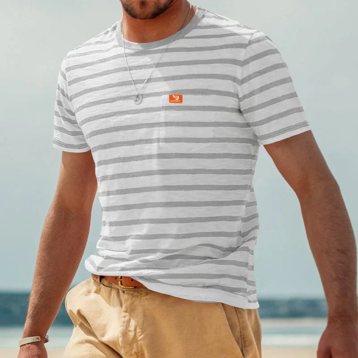 Man wearing gray and white striped T-shirt with small orange logo, beige shorts, and silver necklace at the beach