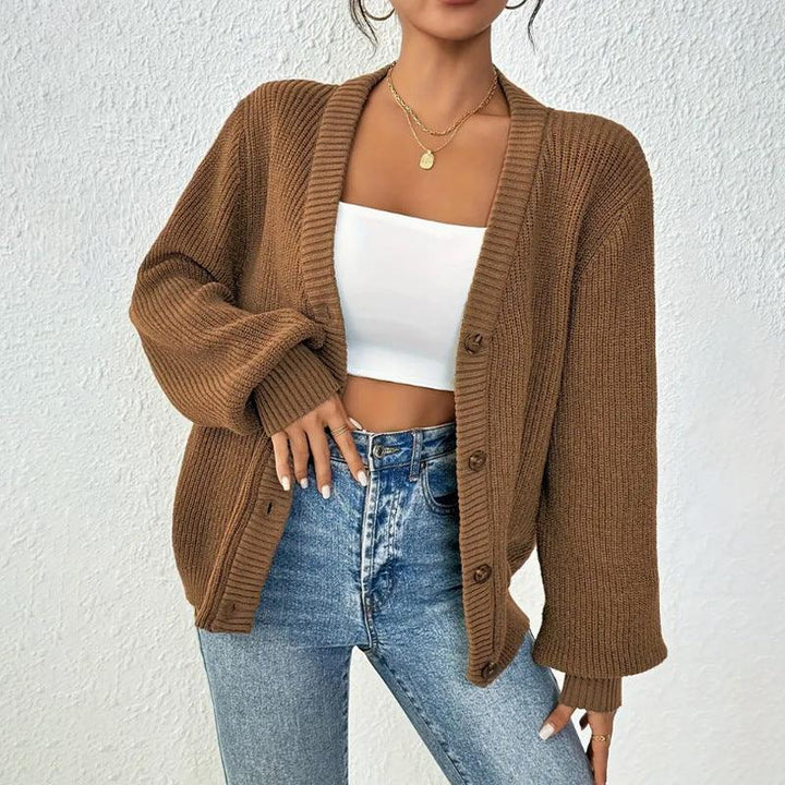 Woman wearing brown button-up knit cardigan over white crop top and high-waisted jeans against white wall