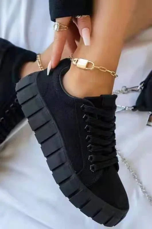 Black chunky platform sneaker with thick sole and black laces, worn with gold chain anklet and manicured nails