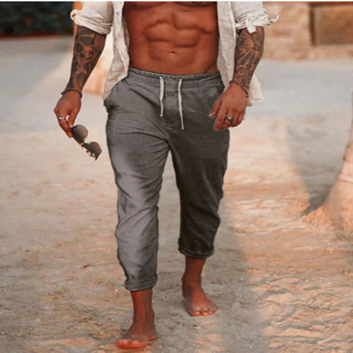 Muscular tattooed man walking barefoot on sandy beach wearing gray jogger pants and holding sunglasses