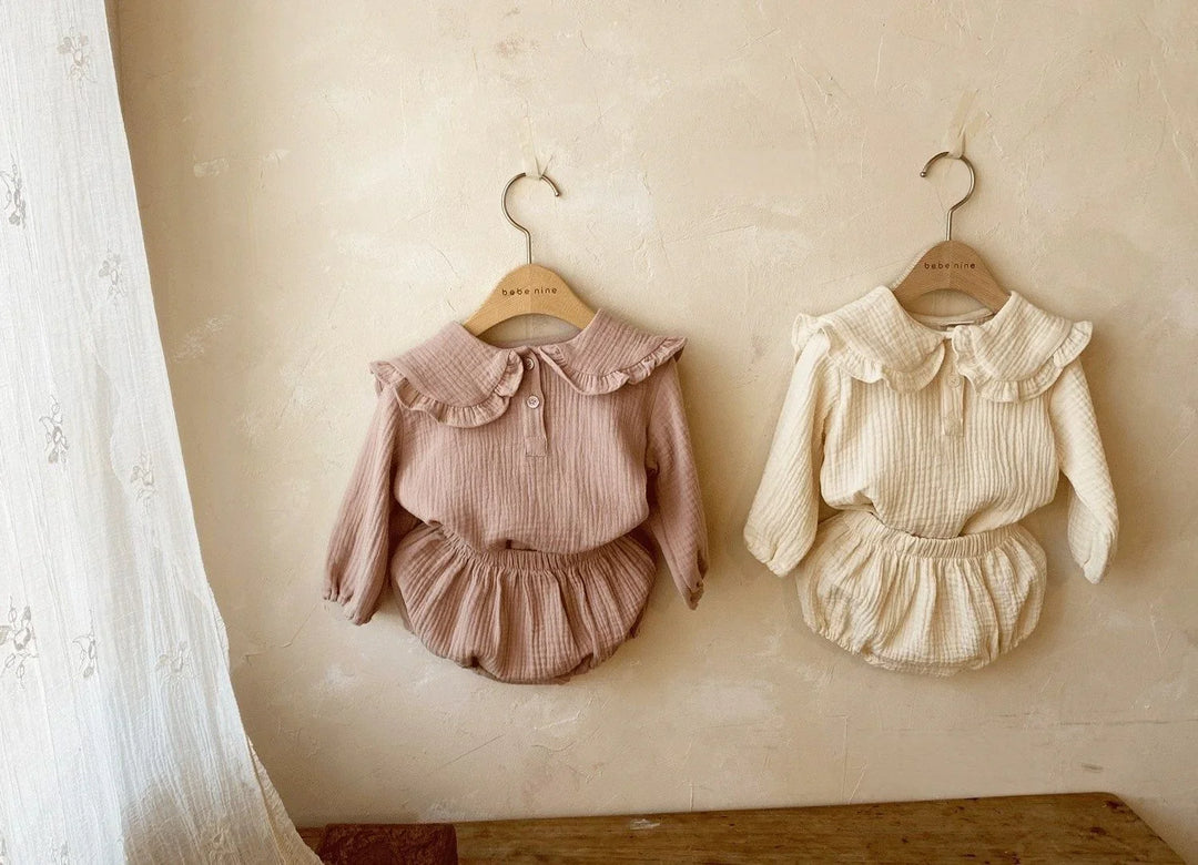 Two baby long-sleeve romper outfits with ruffled collars hanging on beige wall