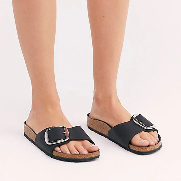 Feet wearing black slide sandals with large silver buckles and cork soles on white background
