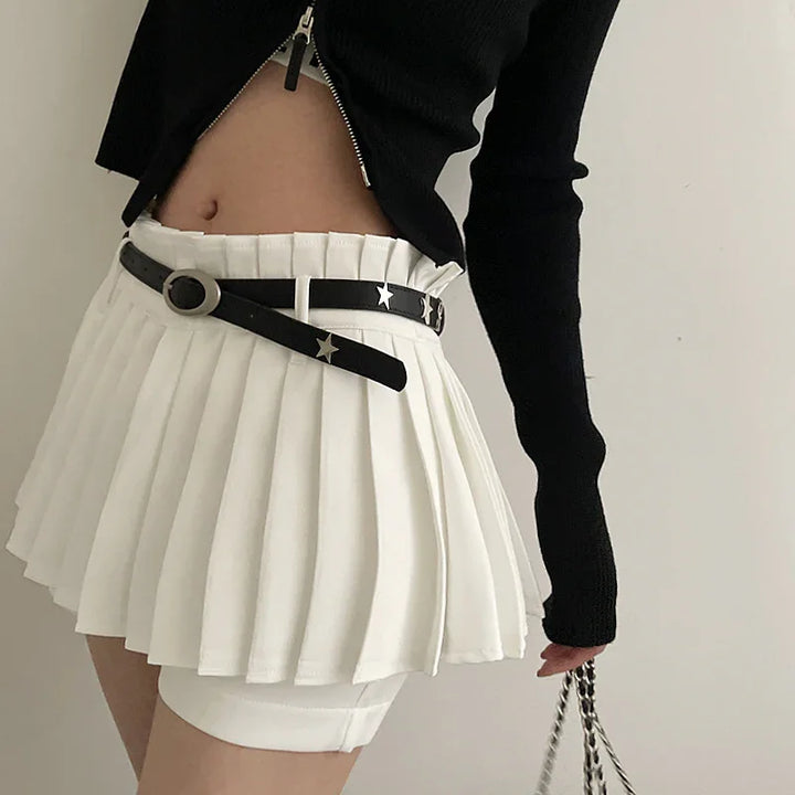 Woman wearing white pleated mini skirt with black star belt and black cropped top holding chain bag