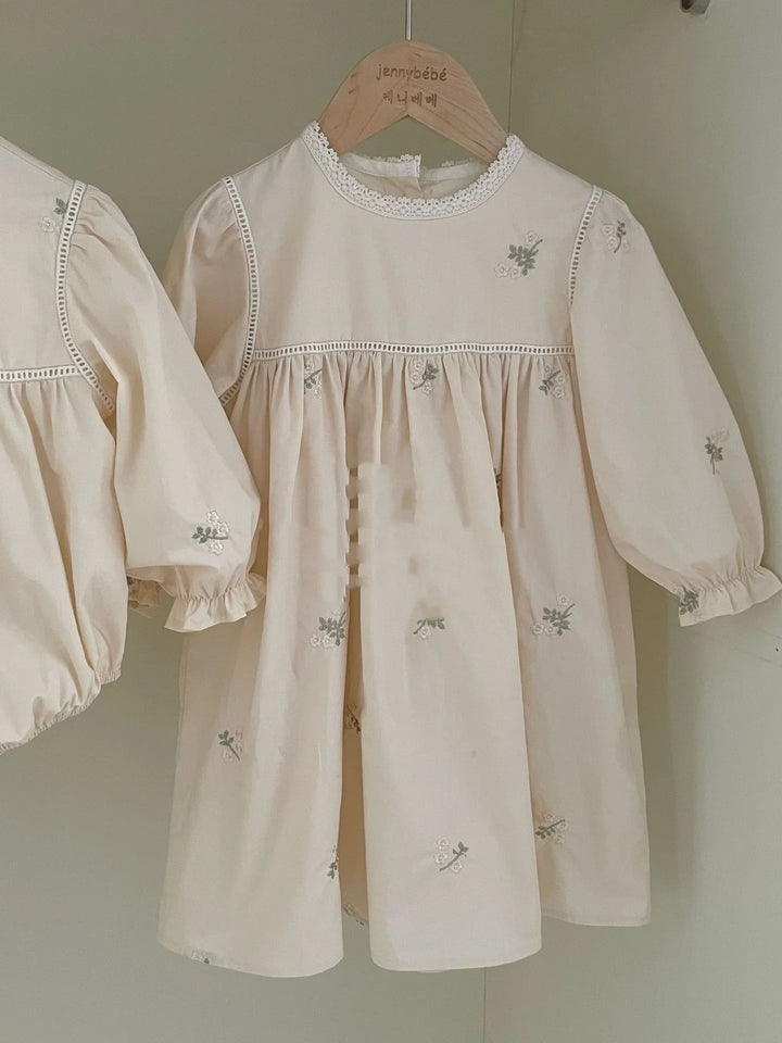 Cream-colored baby dress with floral embroidery and lace details on wooden hanger