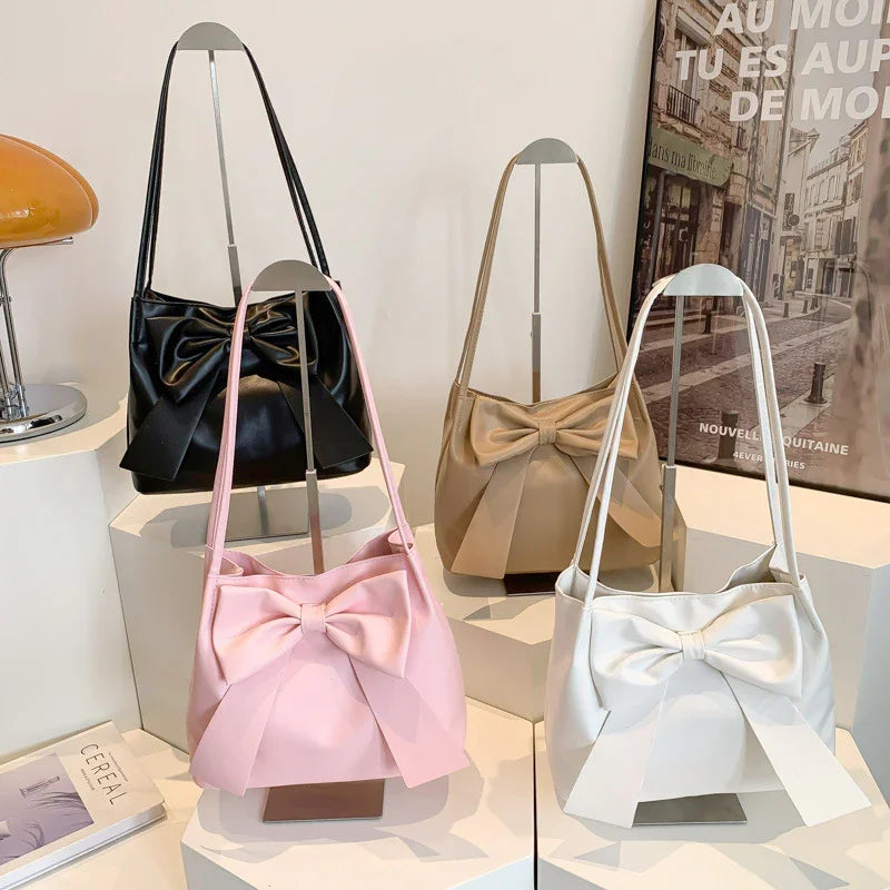 Four stylish leather shoulder bags with large bows in black, beige, pink, and white displayed on white stands